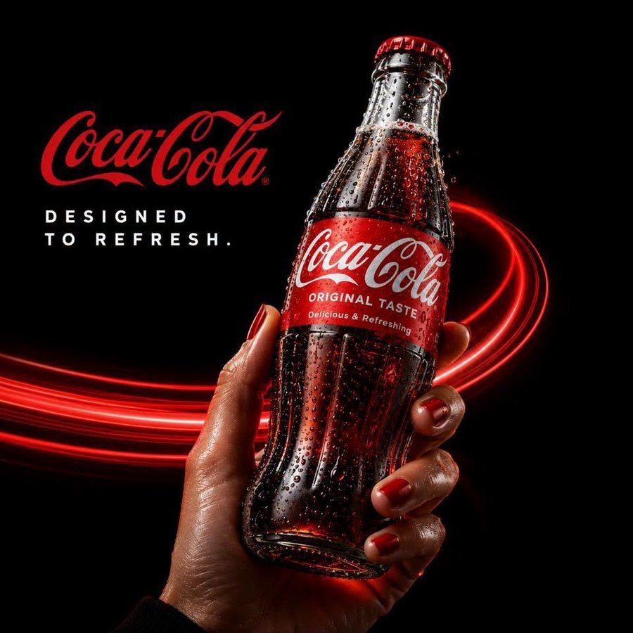Coca-Cola DESIGNED TO REFRESH website hero — hand holding glass bottle on black background with red light swirls